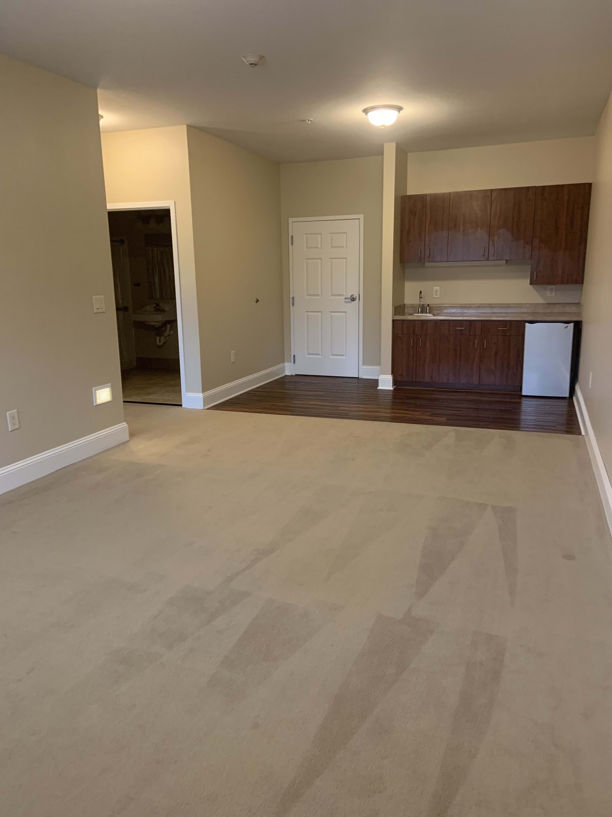 An unfurnished apartment space with a kitchenette and carpeted flooring.