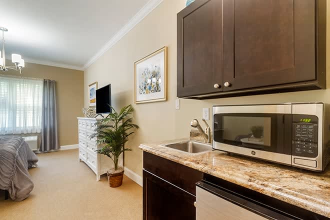 A cozy kitchenette area with a microwave, sink, dark cabinets, and a small seating area visible in the background alongside a television and dresser.
