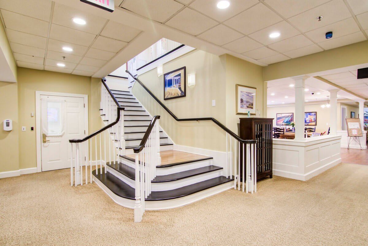 A well-lit lobby featuring a grand staircase with white railings and framed artwork on the walls.