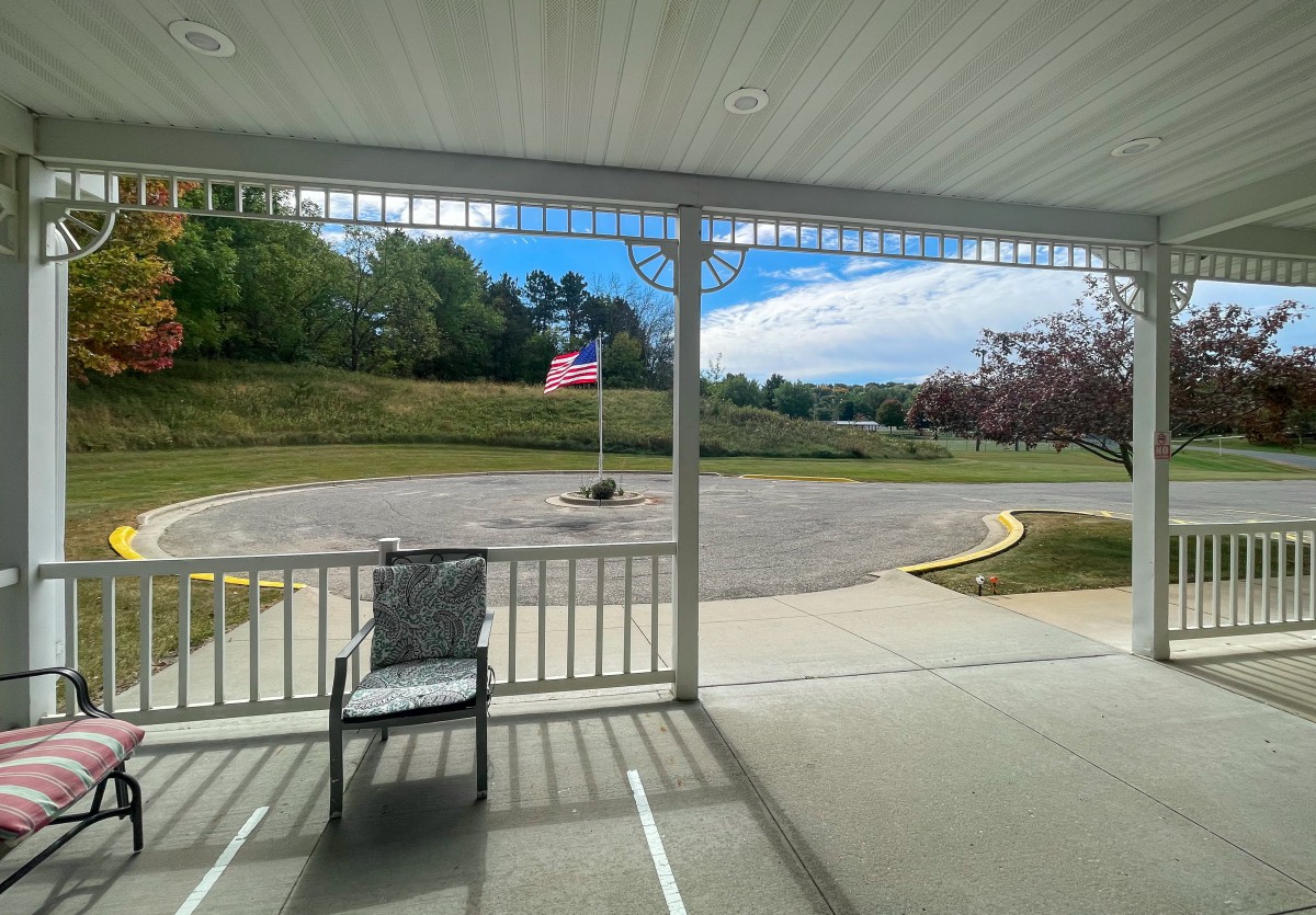 A peaceful view from a porch overlooking an American flag and a quiet road with trees in the background.