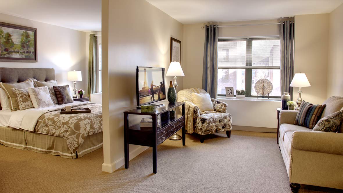 A cozy interior featuring a living area with a TV, comfortable chair, and lamp, adjacent to a bedroom.