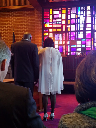 A couple participating in a ceremony, viewed from behind, with vibrant stained glass in the background.
