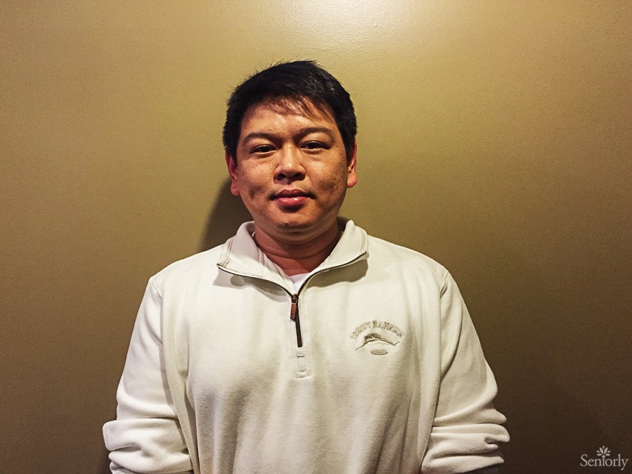 A man wearing a white sweater stands against a plain wall.