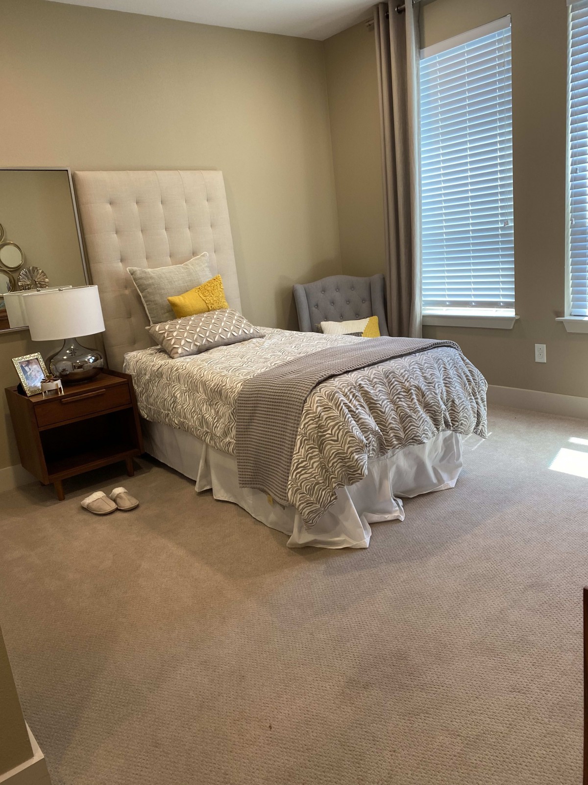 A cozy bedroom featuring a single bed with decorative pillows and a blanket, accompanied by a nightstand, lamp, and armchair near large windows with blinds.