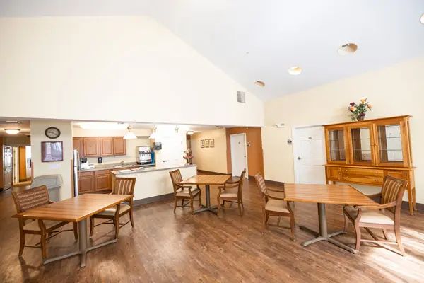 A spacious dining area with wooden floors, featuring several tables and chairs adjacent to an open kitchen.
