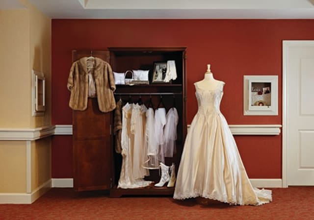 A vintage wedding dress on a mannequin beside an open wardrobe displaying various pieces of attire, including a fur coat and boots.