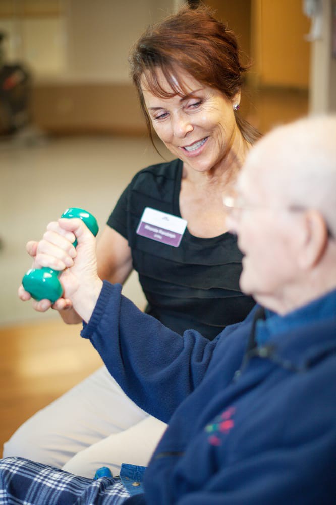 A caregiver assists an elderly person with exercise using a dumbbell.