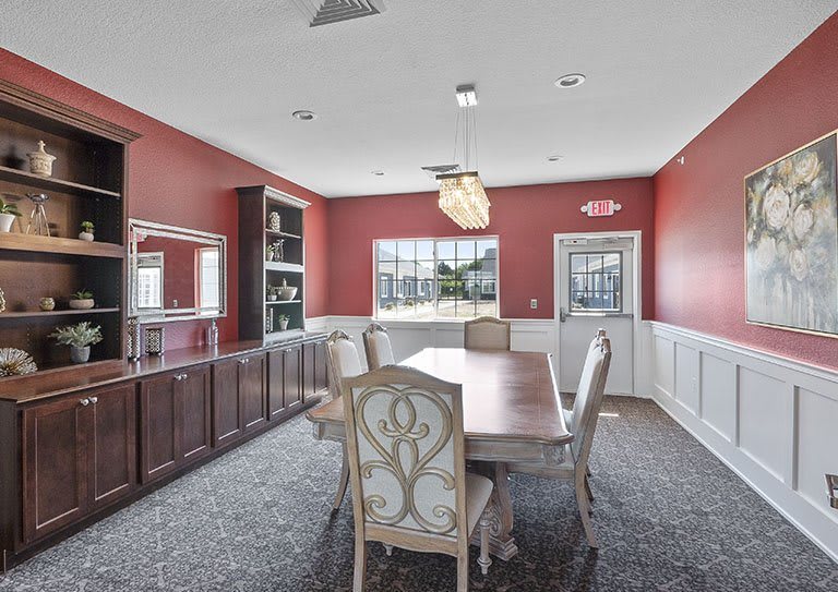 A spacious dining room with red walls, a wooden table, and elegant chairs, featuring a large window and built-in bookshelf.