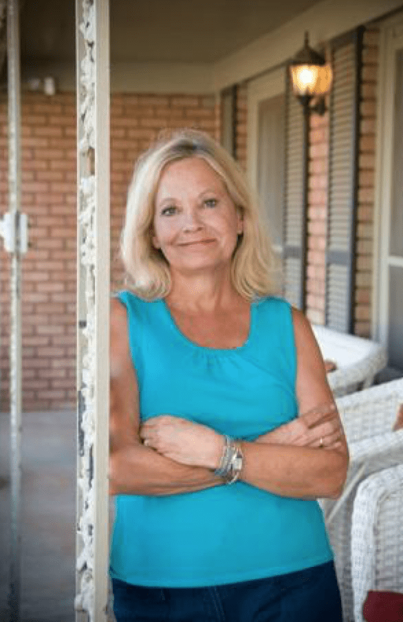 A smiling woman with blonde hair stands on a porch, wearing a blue shirt with her arms crossed.