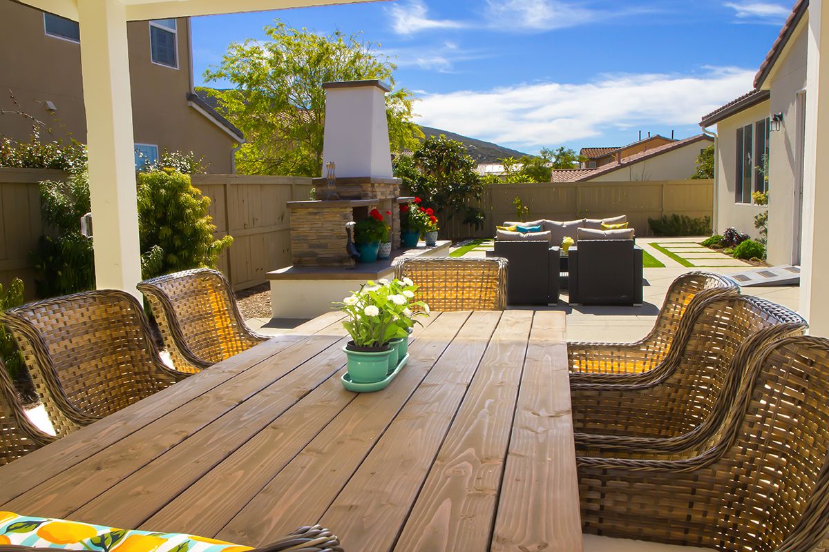 A cozy backyard patio with a wooden dining table, chairs, and an outdoor fireplace under a bright blue sky.