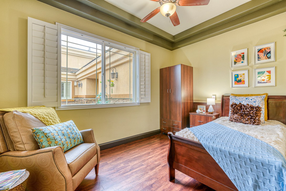 A cozy bedroom with wooden flooring, a ceiling fan, and ample natural light streaming through the window.