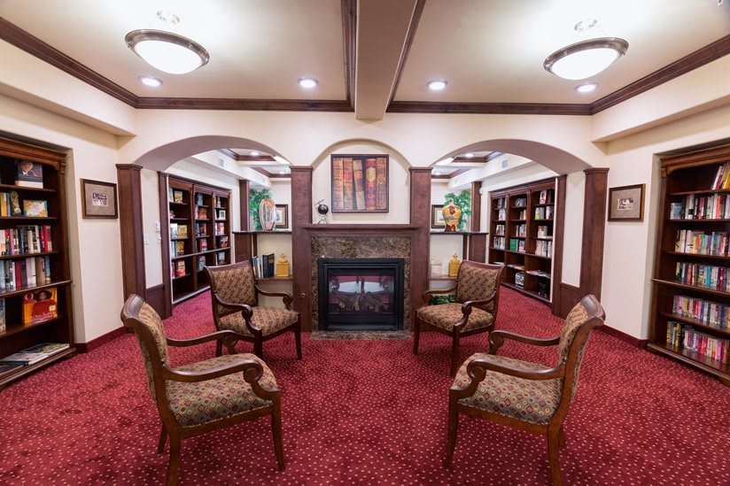 A cozy library interior with chairs arranged around a fireplace, surrounded by bookshelves.
