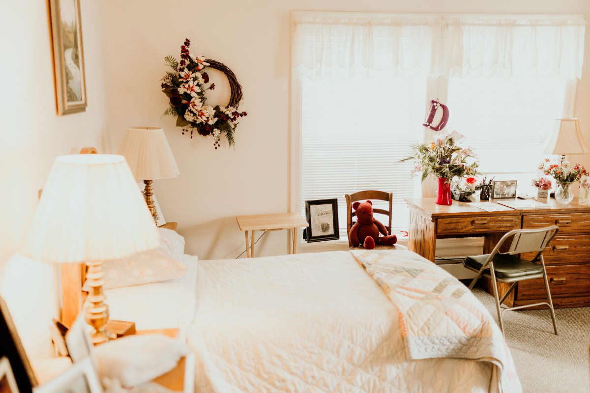 A cozy bedroom with a neatly made bed, a wall wreath, and a desk adorned with flowers.