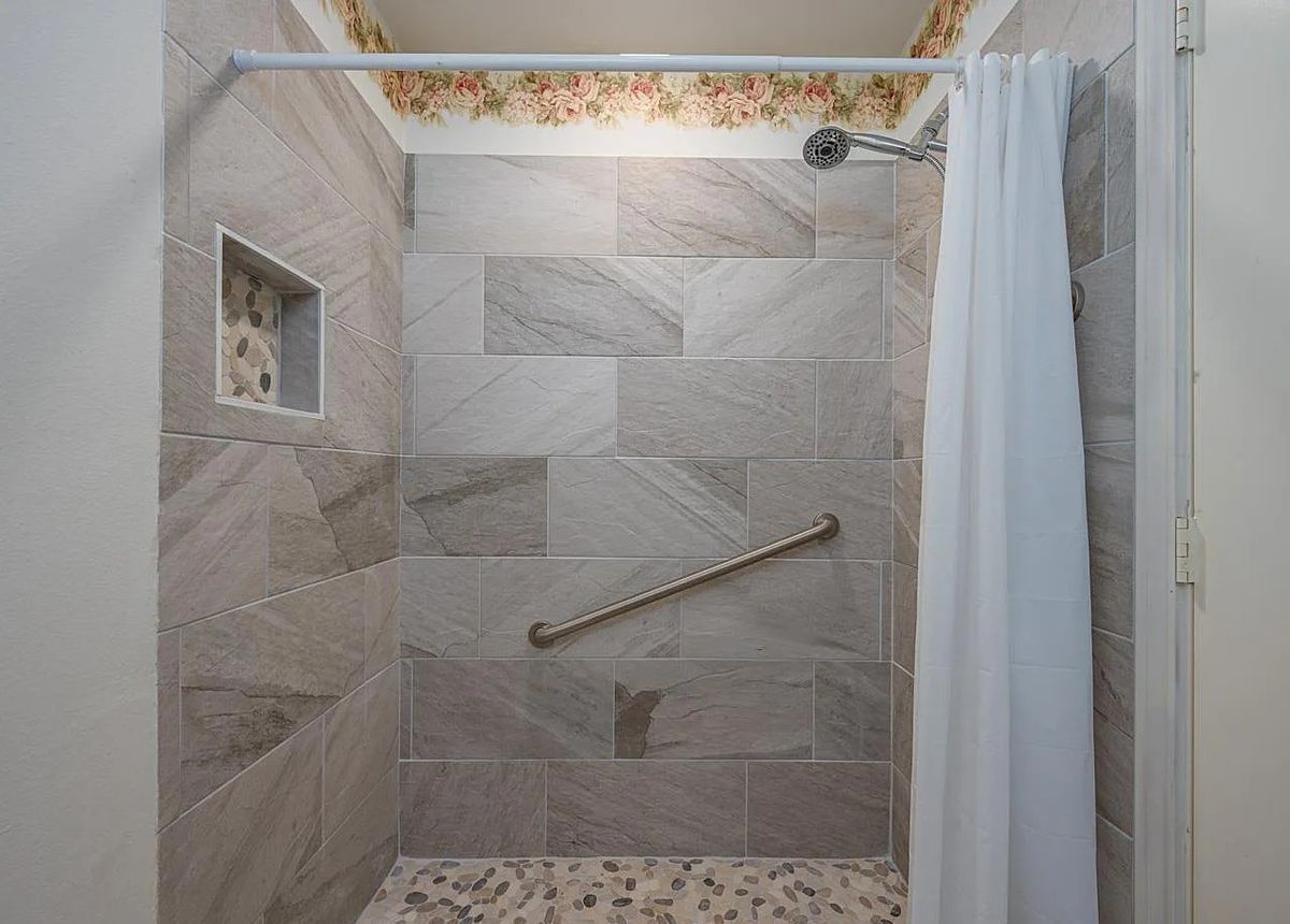 A modern shower with large tiles, a grab bar, and a white shower curtain.