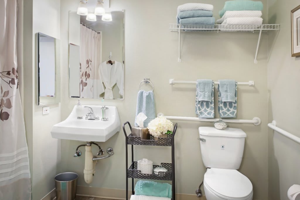 A tidy, minimalist bathroom with a white sink, toilet, and organized storage using an organizer cart and wall shelves. Light blue towels add a touch of color.