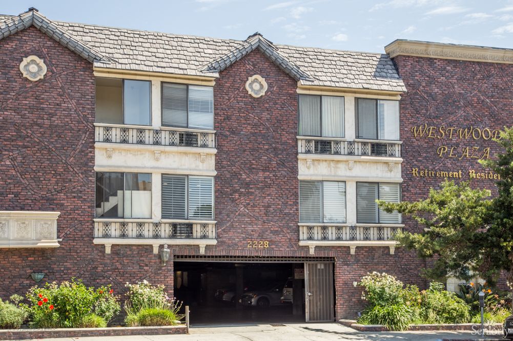 A three-story brick apartment building with large windows and a garage entrance, surrounded by landscaped greenery.