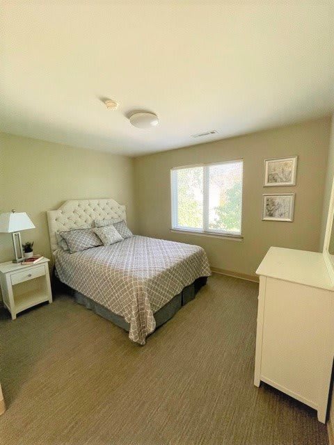 A cozy bedroom with light walls, featuring a neatly made bed, a bedside lamp, and a dresser.