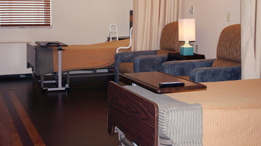 A view of a patient room in a hospital, featuring two beds, a lamp, and seating.