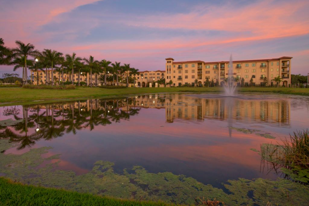 A serene view of a pond reflecting a building and palm trees under a colorful sunset sky.