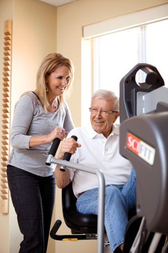 A caregiver assists an elderly man using exercise equipment.