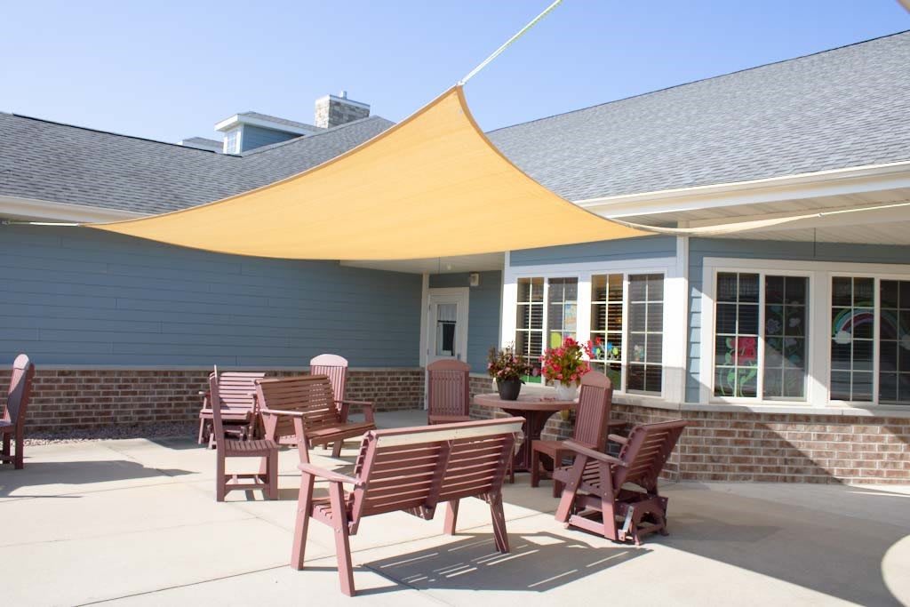 An outdoor patio area with a yellow shade sail, chairs, and a table in front of a building.