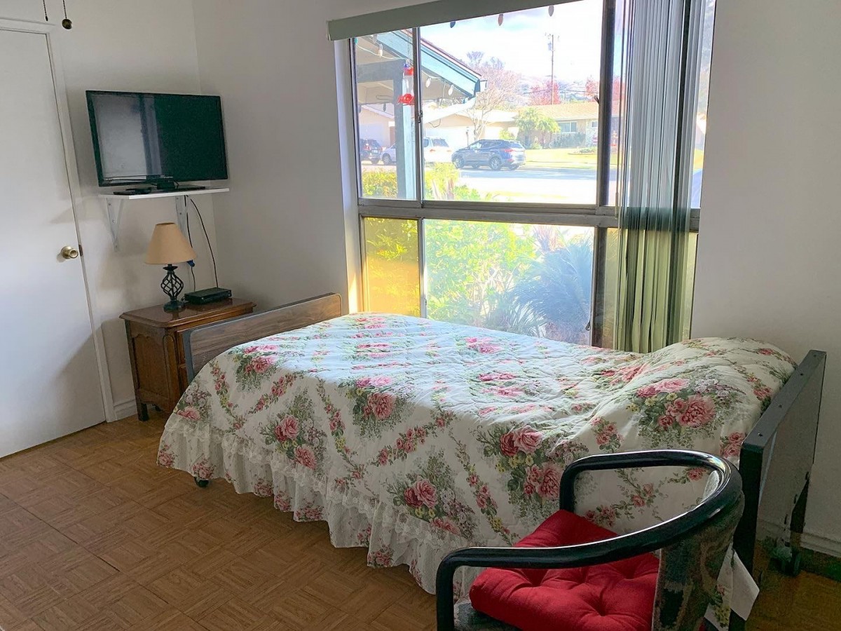 A cozy bedroom with a floral bedspread, a television mounted on the wall, and a large window letting in natural light.