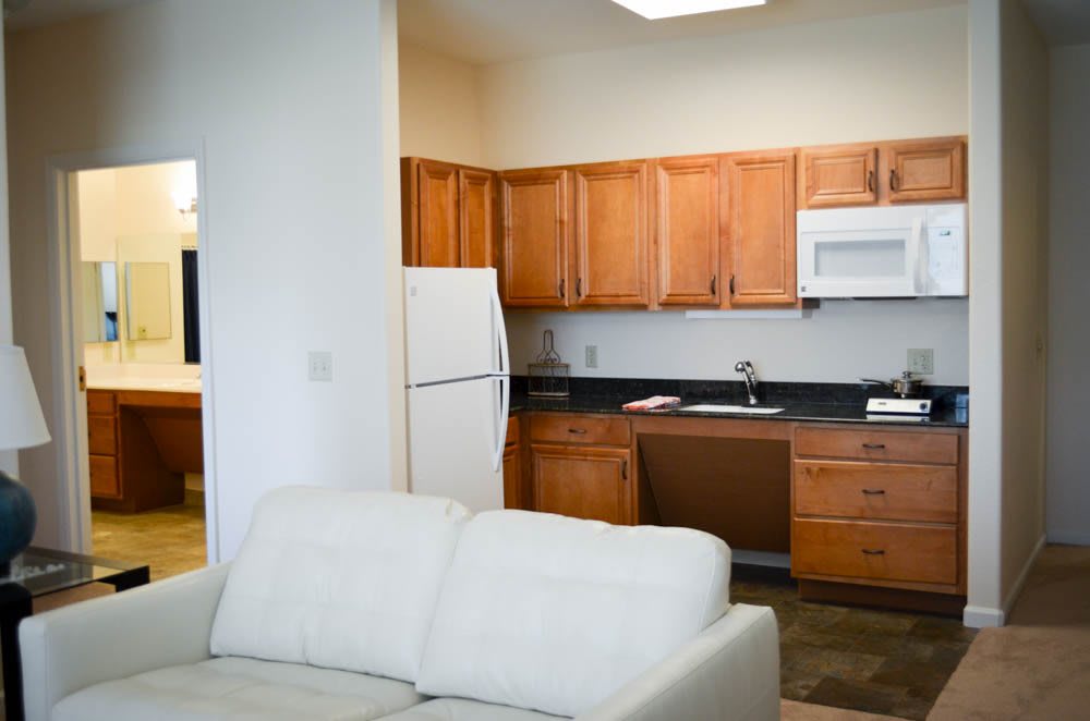 A cozy open-plan kitchen and living area featuring wooden cabinets, a refrigerator, microwave, and a comfortable white sofa.