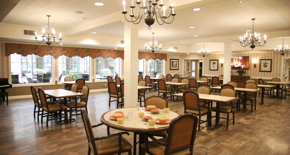 A well-lit dining room with elegant chandeliers, featuring multiple tables and chairs arranged neatly.