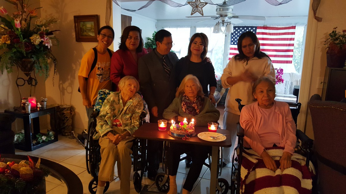 A family gathering celebrating a birthday with elderly members in wheelchairs, surrounded by loved ones indoors, with candles and a flag in the background.