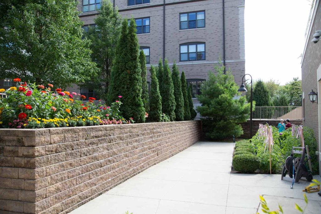 A landscaped outdoor area with a brick wall, flowers, trees, and a sidewalk next to a building.