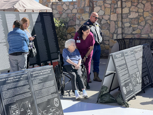 An elderly individual in a wheelchair observes informational displays at an outdoor event, assisted by a caregiver.