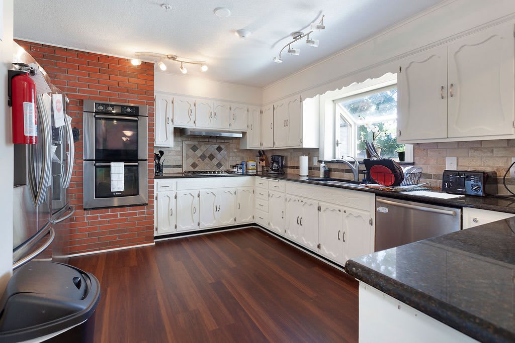 A modern kitchen with wood flooring and white cabinets, featuring a brick accent wall and granite countertops.