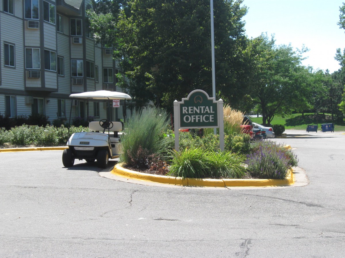 A rental office sign located near an apartment complex, with a golf cart parked nearby and landscaping in the surroundings.