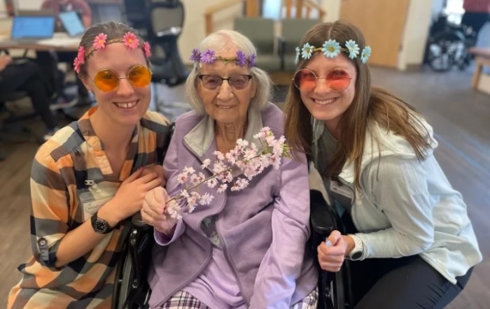 Three women, one elderly and in a wheelchair, wearing flower crowns and smiling.