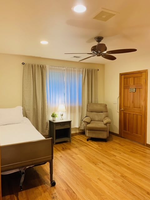 A cozy bedroom with wooden flooring, featuring a bed, armchair, ceiling fan, and a side table with a lamp.