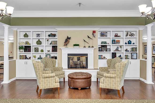 A cozy living room featuring four patterned armchairs arranged around a round coffee table, with a fireplace and decorated shelves in the background.