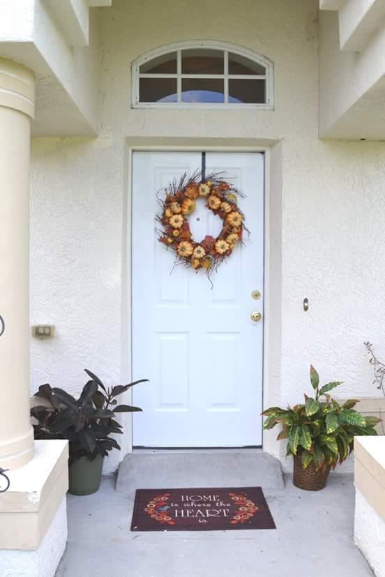 A front door decorated with a sunflower wreath, flanked by potted plants.