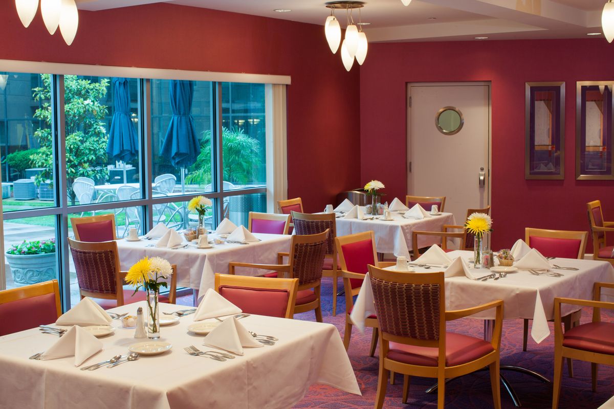 A well-arranged restaurant dining room with neatly set tables and chairs, featuring large windows providing a view of the outside.