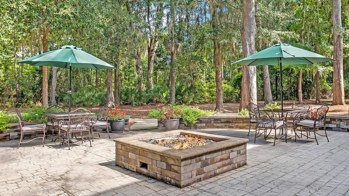 A serene outdoor patio area featuring a central fire pit, surrounded by seating and large umbrellas, nestled in a forested setting.