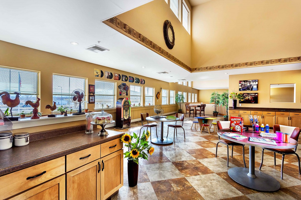 A bright and welcoming dining area with tables, chairs, and plants, illuminated by natural sunlight through large windows.
