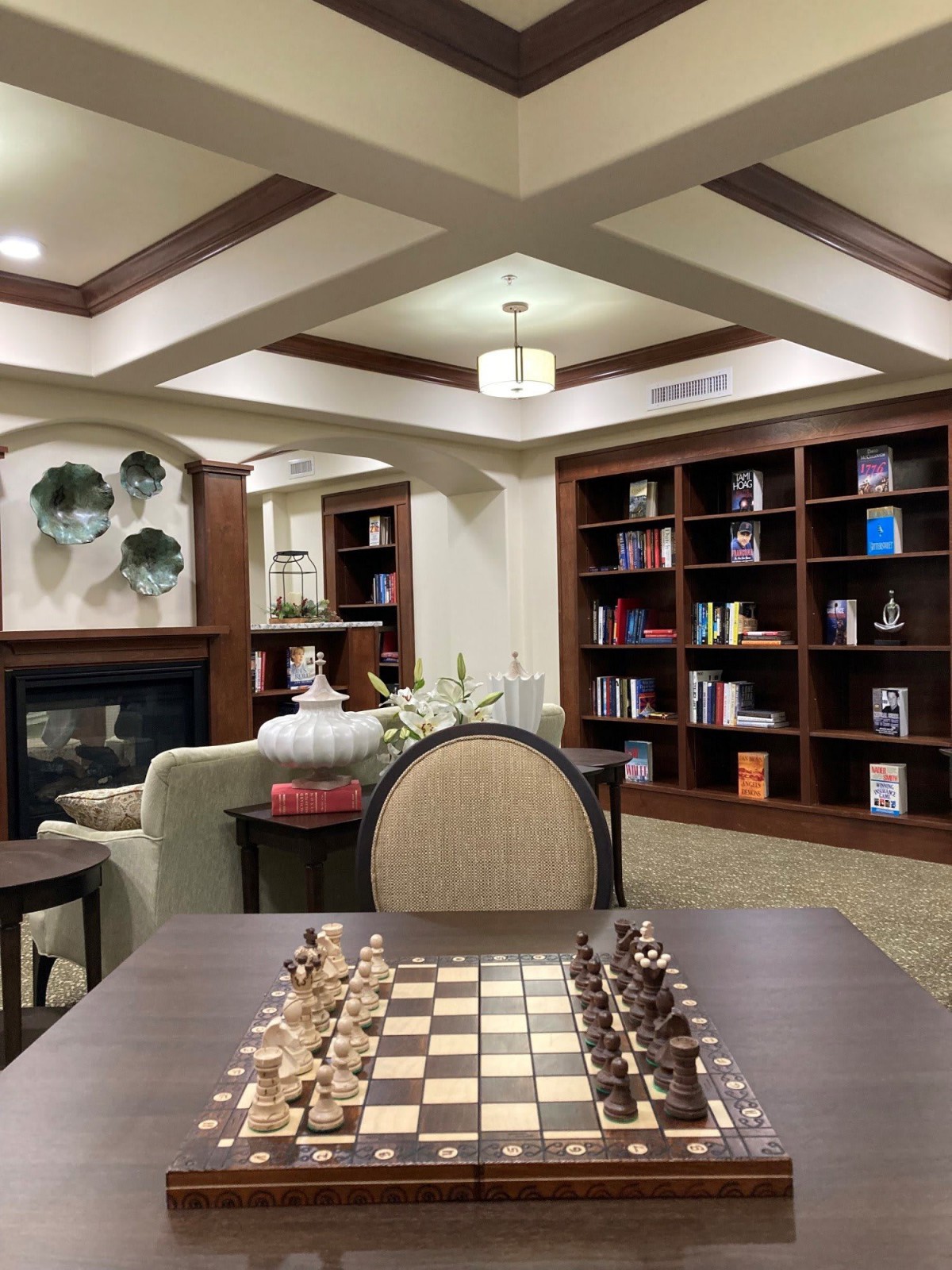 A cozy library interior with bookshelves, a fireplace, and a chess board on the table.