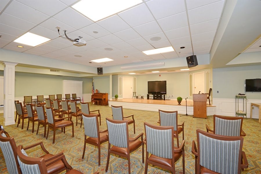 A spacious conference room with a seating arrangement facing a stage equipped with a podium and a large TV screen.