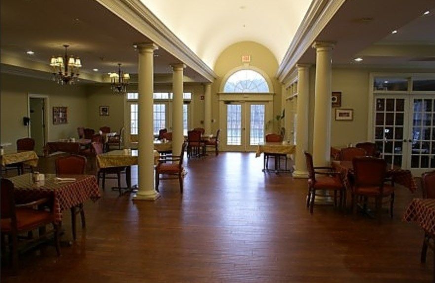 An empty dining area with wooden floors, tables, and chairs lined along a columned hallway.