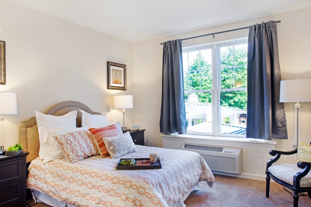 A cozy bedroom with a large window letting in natural light, featuring decorative pillows and a stylish bedspread.