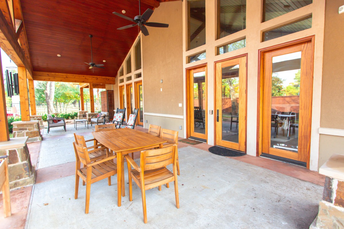 An outdoor patio area with wooden dining furniture and ceiling fans, adjacent to a building with large glass doors.