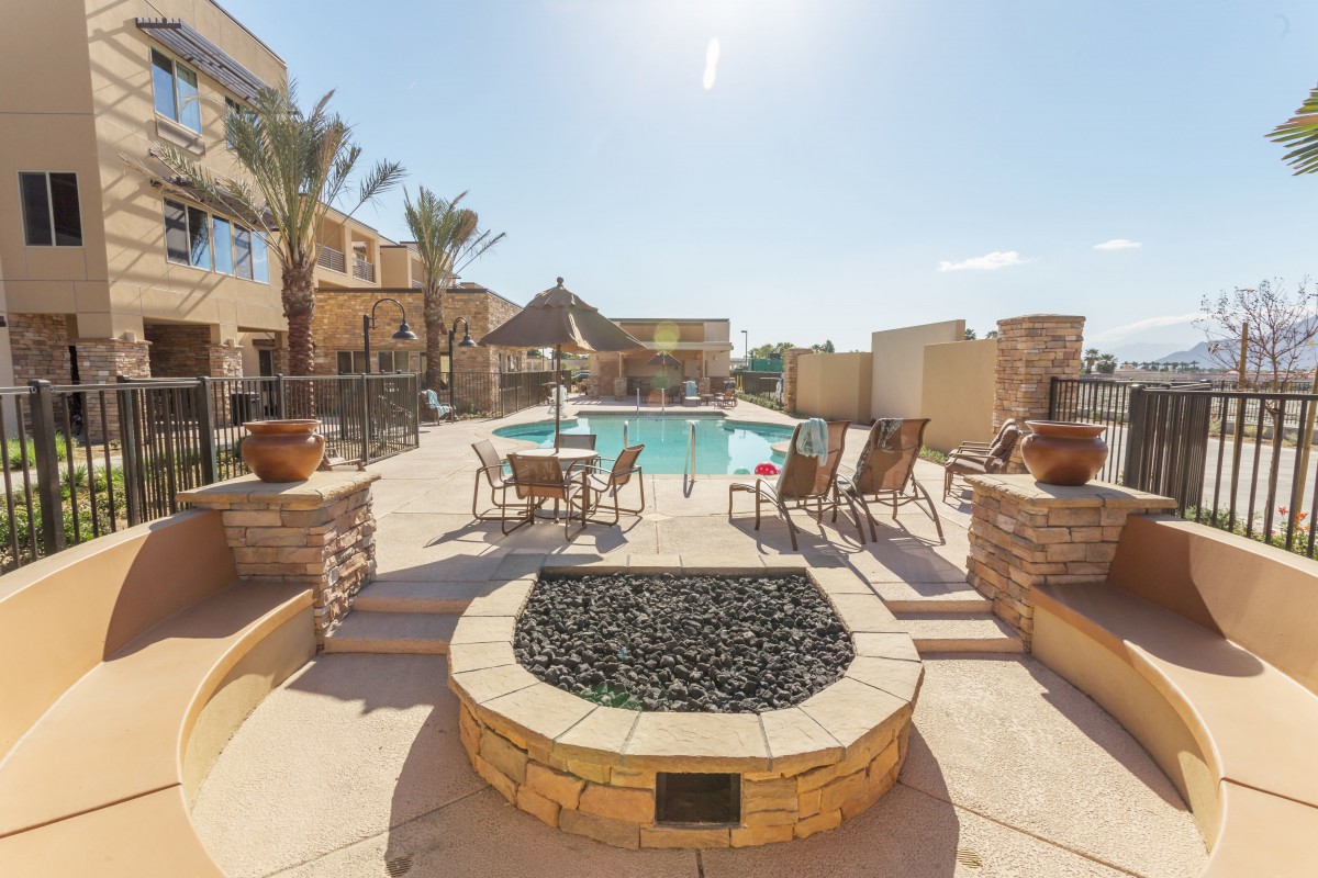 A sunny outdoor pool area with lounge chairs and a fire pit surrounded by palm trees.