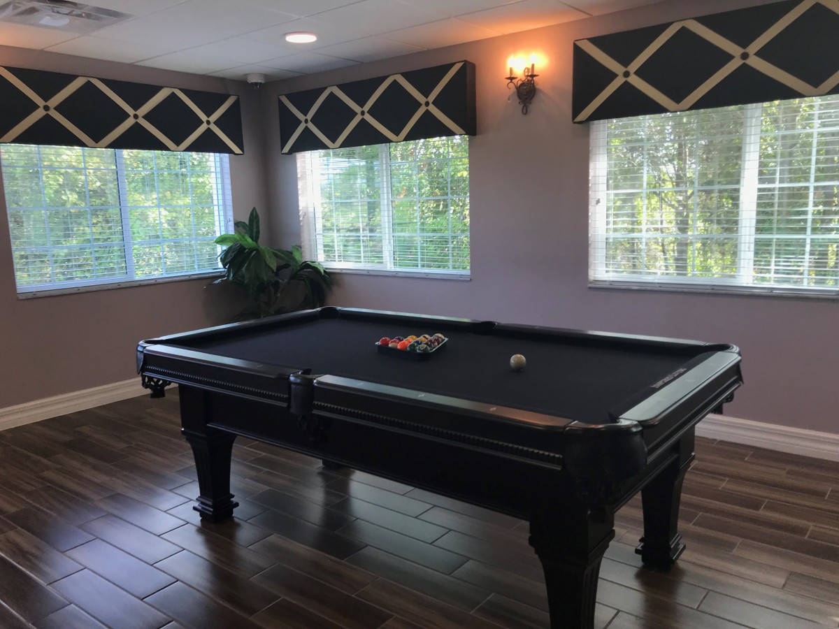 A stylish recreation room featuring a black pool table with elegant window treatments and hardwood flooring.
