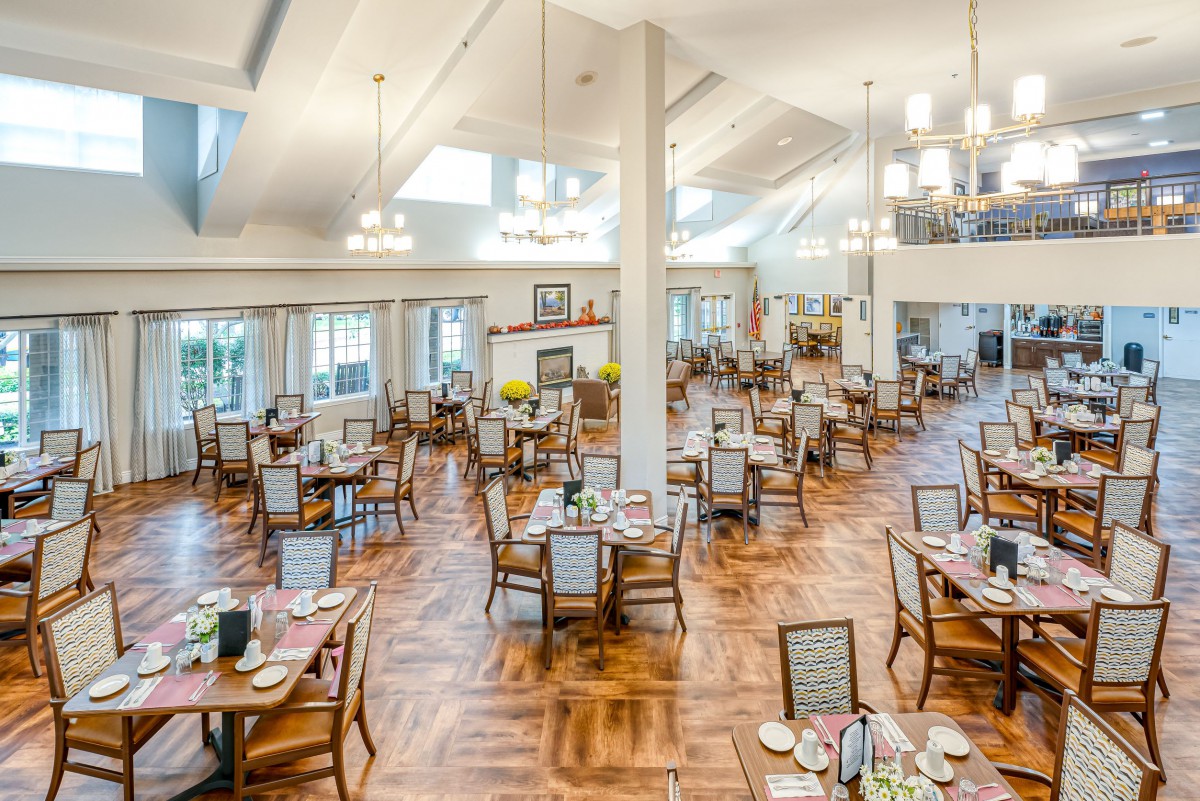 A spacious and elegantly decorated dining area with multiple tables and chairs, featuring a wooden floor and bright lighting from chandeliers.