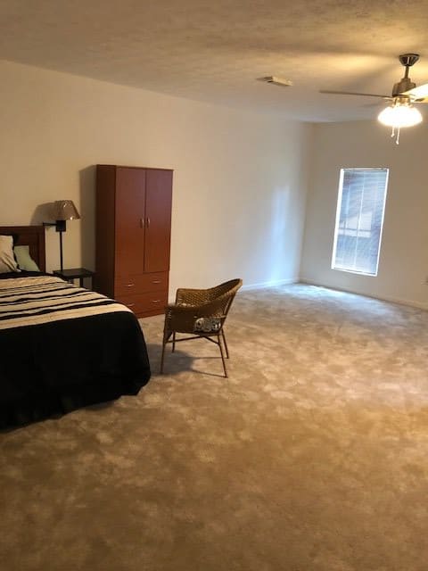 A spacious bedroom with a bed, wardrobe, and chair on a carpeted floor, featuring a window and ceiling fan.