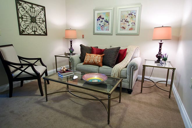 A cozy living room with a sofa and decorative pillows, flanked by side tables and lamps, featuring artwork on the walls.
